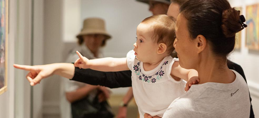 Photograph of woman holding a baby that is pointing at art